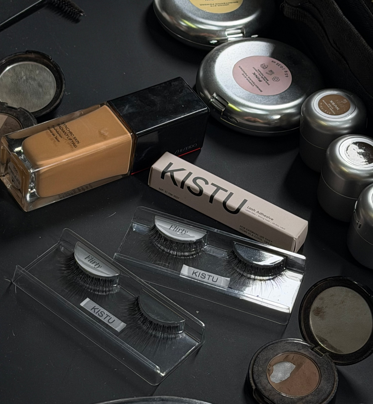 Makeup products including eyelashes and compact powders on a black surface with 'KISTU' branding.